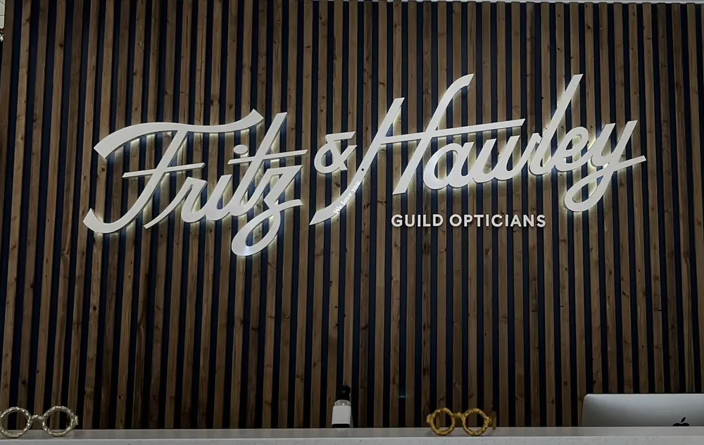 The Fritz & Hawley Guild Opticians name in white script lettering across a wall of dark wood slats, with sculptural eyeglass frames set out along the counter below