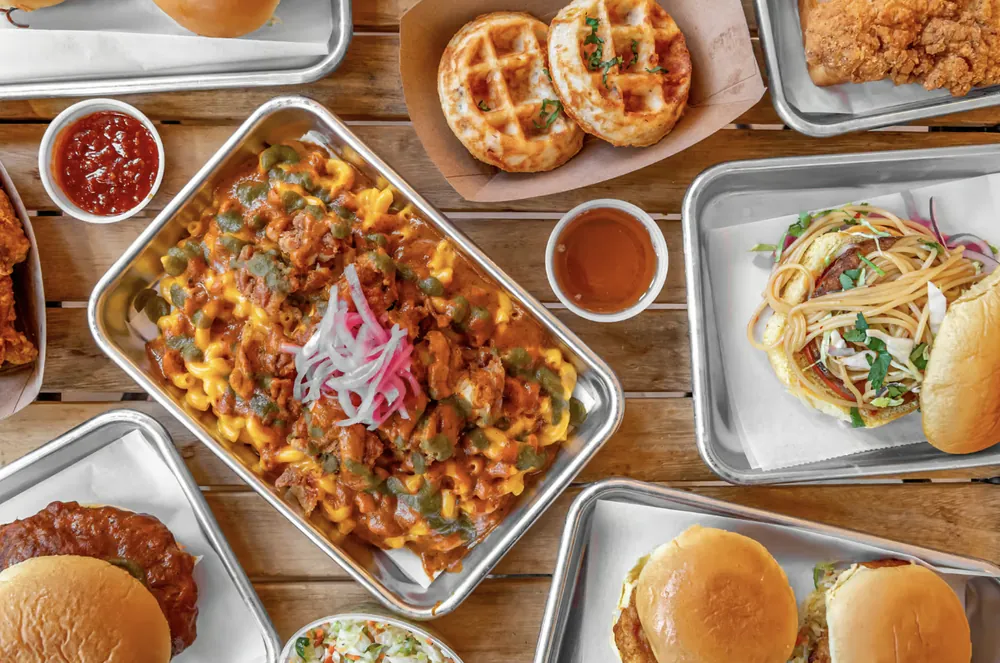 Metal trays of Hot Murga fried chicken, loaded macaroni, chicken and waffles and sandwiches, seen from above on a wooden table