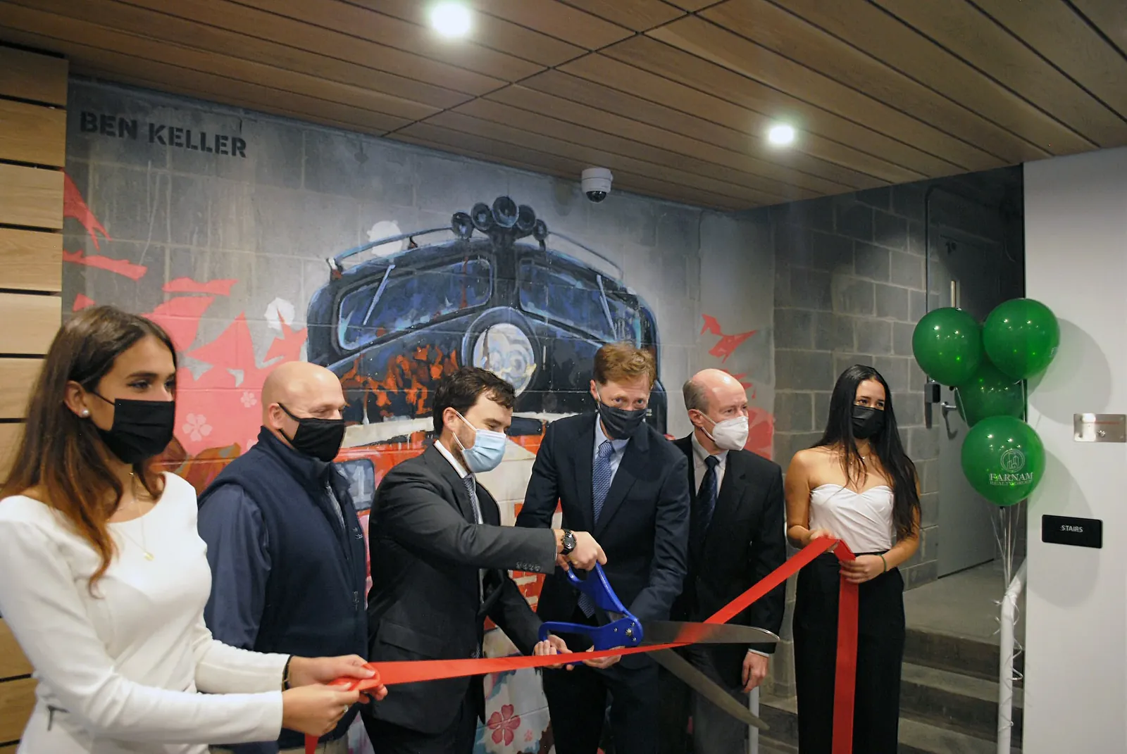 Six people in face masks cutting a red ribbon with oversized ceremonial scissors in the lobby of The Elm, in front of a large mural of a locomotive signed Ben Keller, with green balloons alongside.