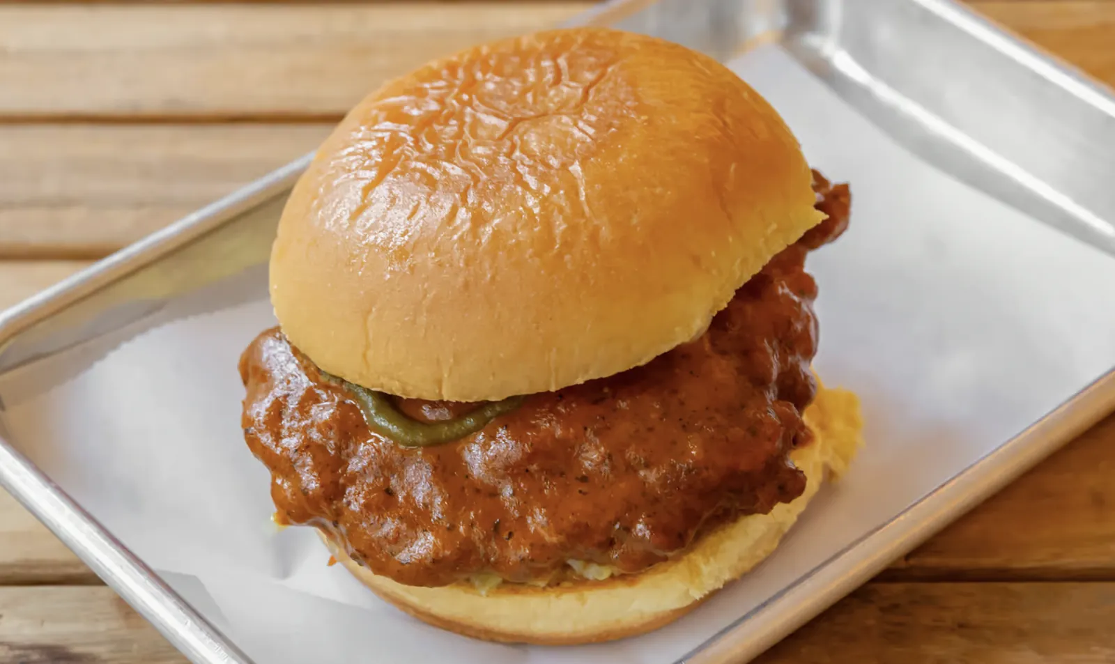 A fried chicken sandwich from Hot Murga, coated in orange spiced sauce and topped with green chillies, served in a glazed bun on a metal tray.