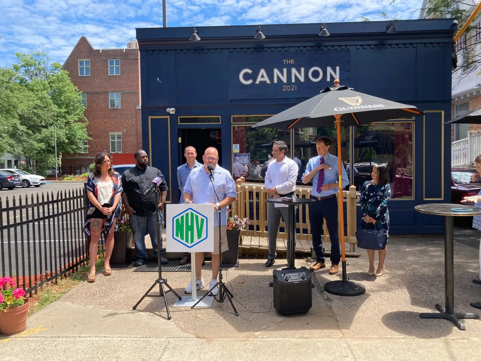 An outdoor press event on the pavement outside The Cannon, a navy blue building lettered 'The Cannon 2021'. A man speaks at a lectern carrying the New Haven NHV logo while six people stand behind him under a patio umbrella.