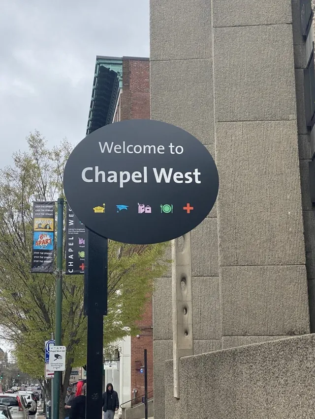 An oval 'Welcome to Chapel West' sign mounted on a post at the edge of the district, carrying the five Chapel West category icons, with a Chapel West street banner on the lamppost behind it.
