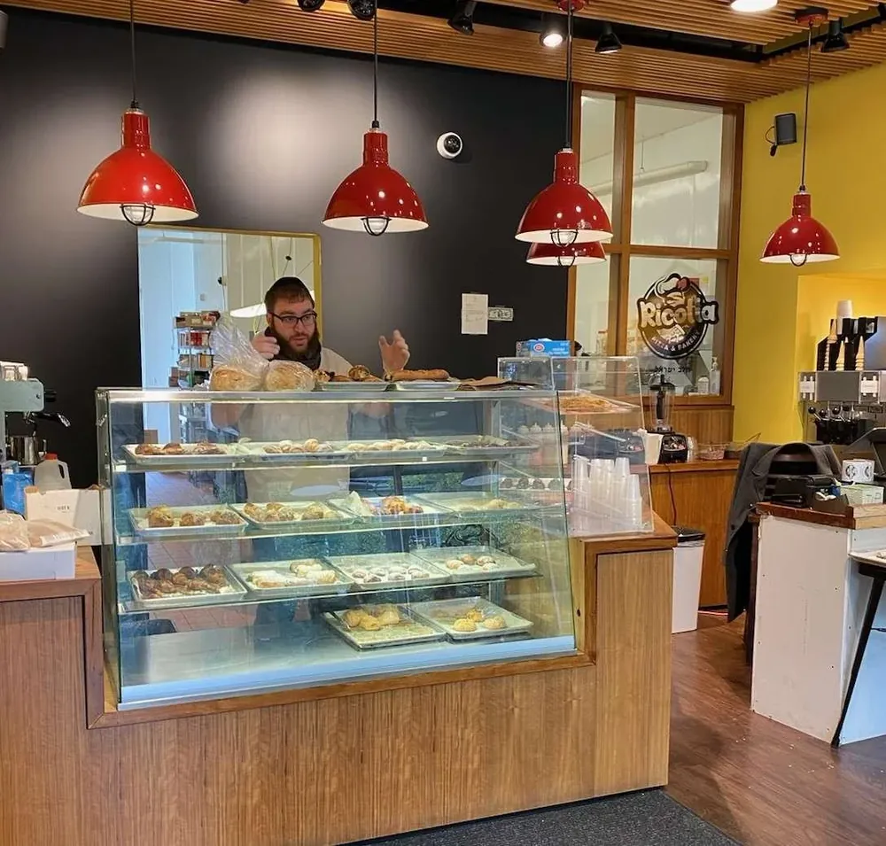 The interior of Ricotta Pizza and Bakery, with a glass case of pastries along the counter under a row of red pendant lamps, and a member of staff working behind it.
