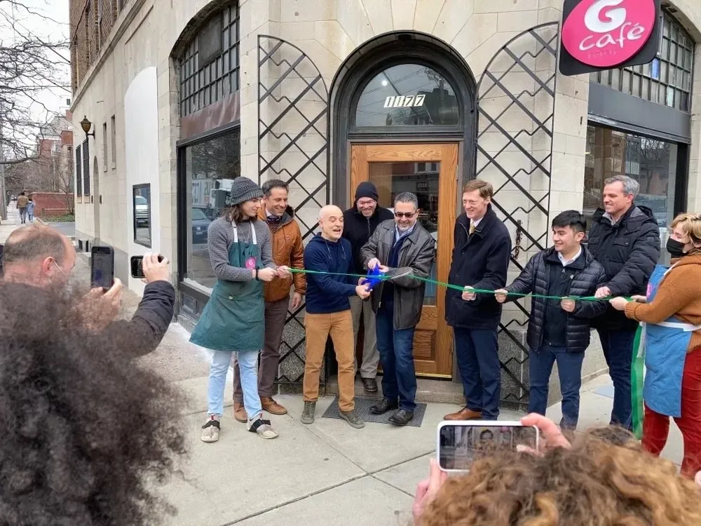 Seven people cutting a green ribbon outside G Cafe Bakery's arched doorway on a winter afternoon, watched by onlookers filming on their phones.