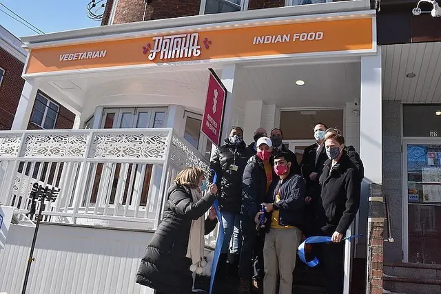 A masked group cutting a blue ribbon on the front steps of Pataka Vegetarian Indian Food, under the restaurant's orange and white sign.