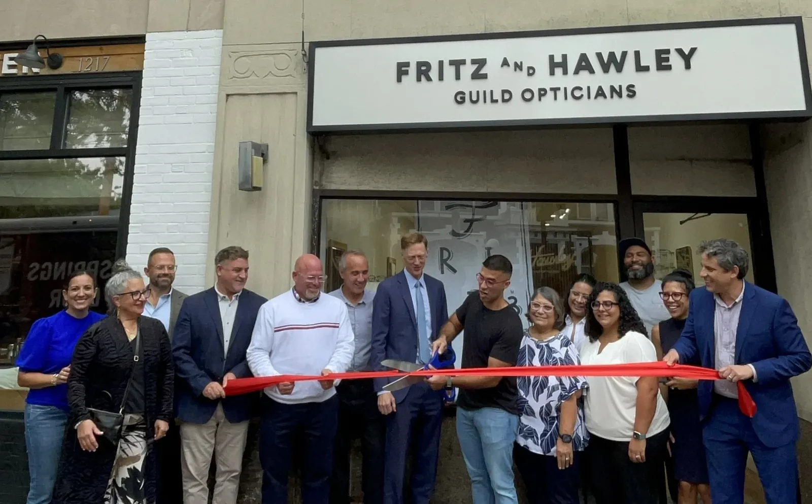 About fourteen people gathered across the pavement to cut a red ribbon outside Fritz and Hawley Guild Opticians, whose name is lettered on the shopfront above them.