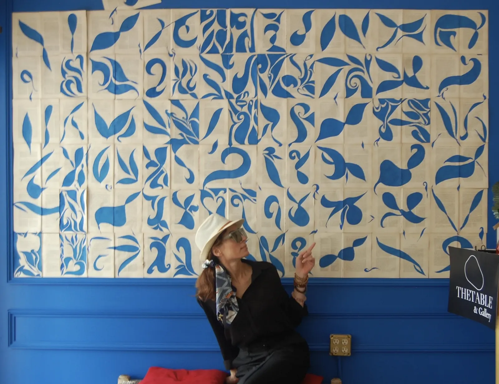A woman in a straw hat sitting beneath a large mural of blue botanical shapes painted across cream panels, pointing up at it, beside a sign reading The Table and Gallery.