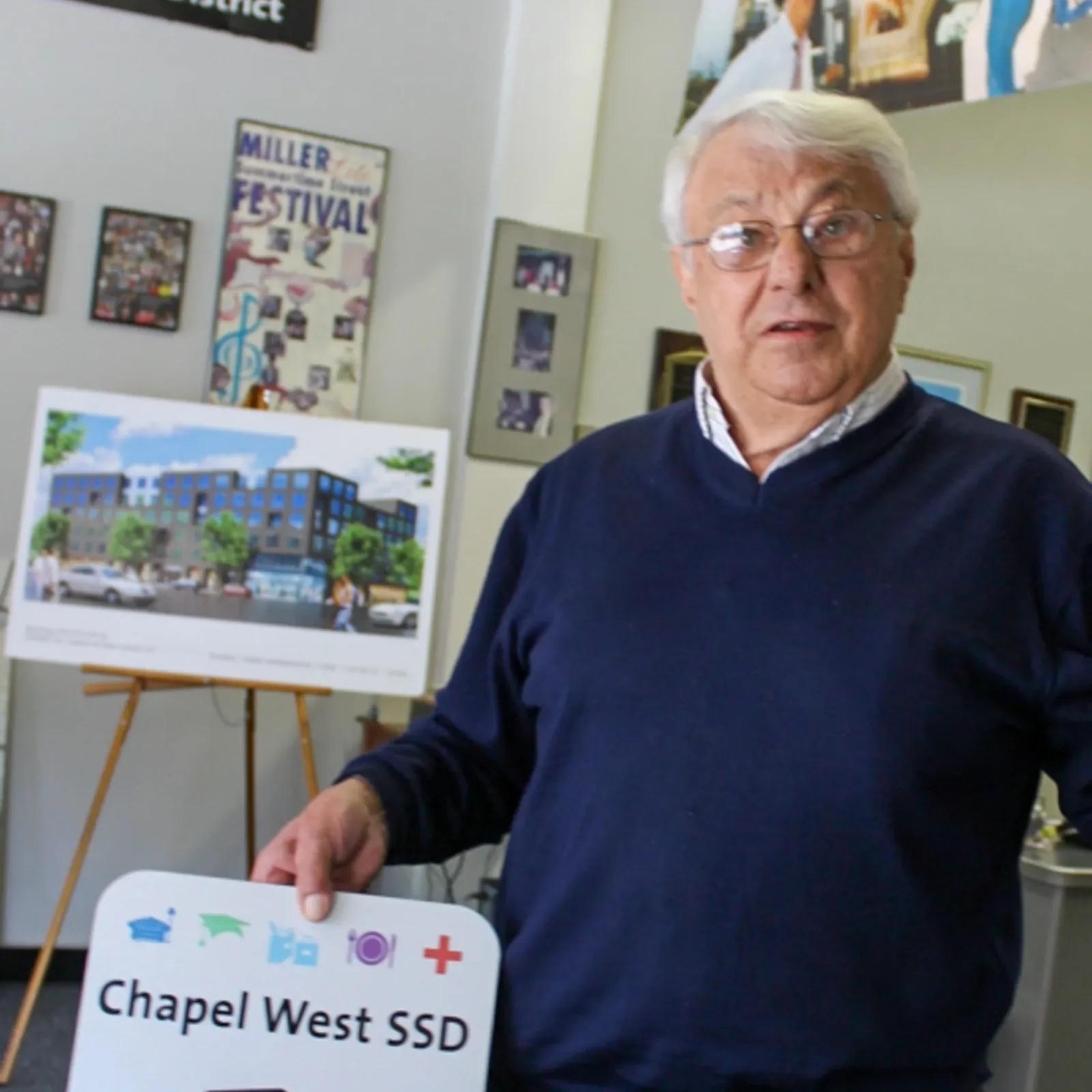 A silver-haired man in a navy sweater standing in the Chapel West district office, resting a hand on a Chapel West SSD sign, with an architectural rendering of a new district building on an easel behind him.
