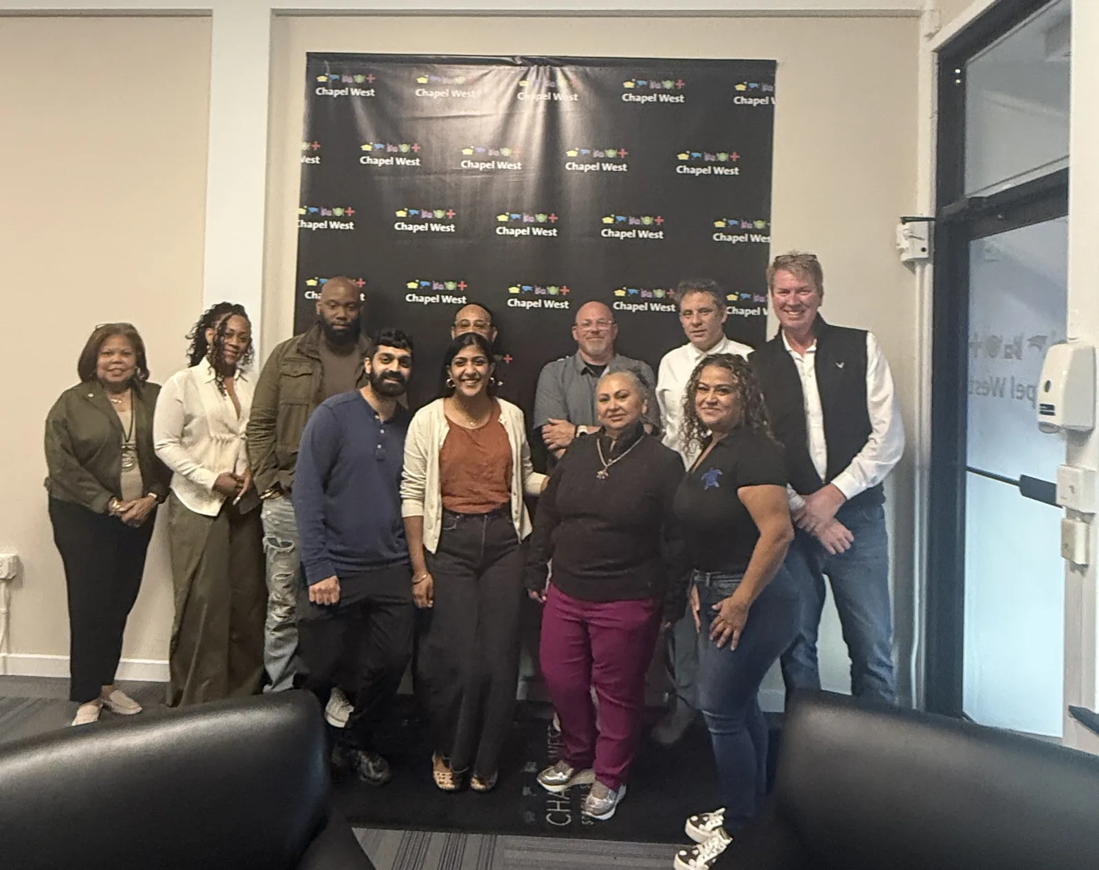 Thirteen business owners and City staff standing together in front of the Chapel West logo backdrop at the district office after the session