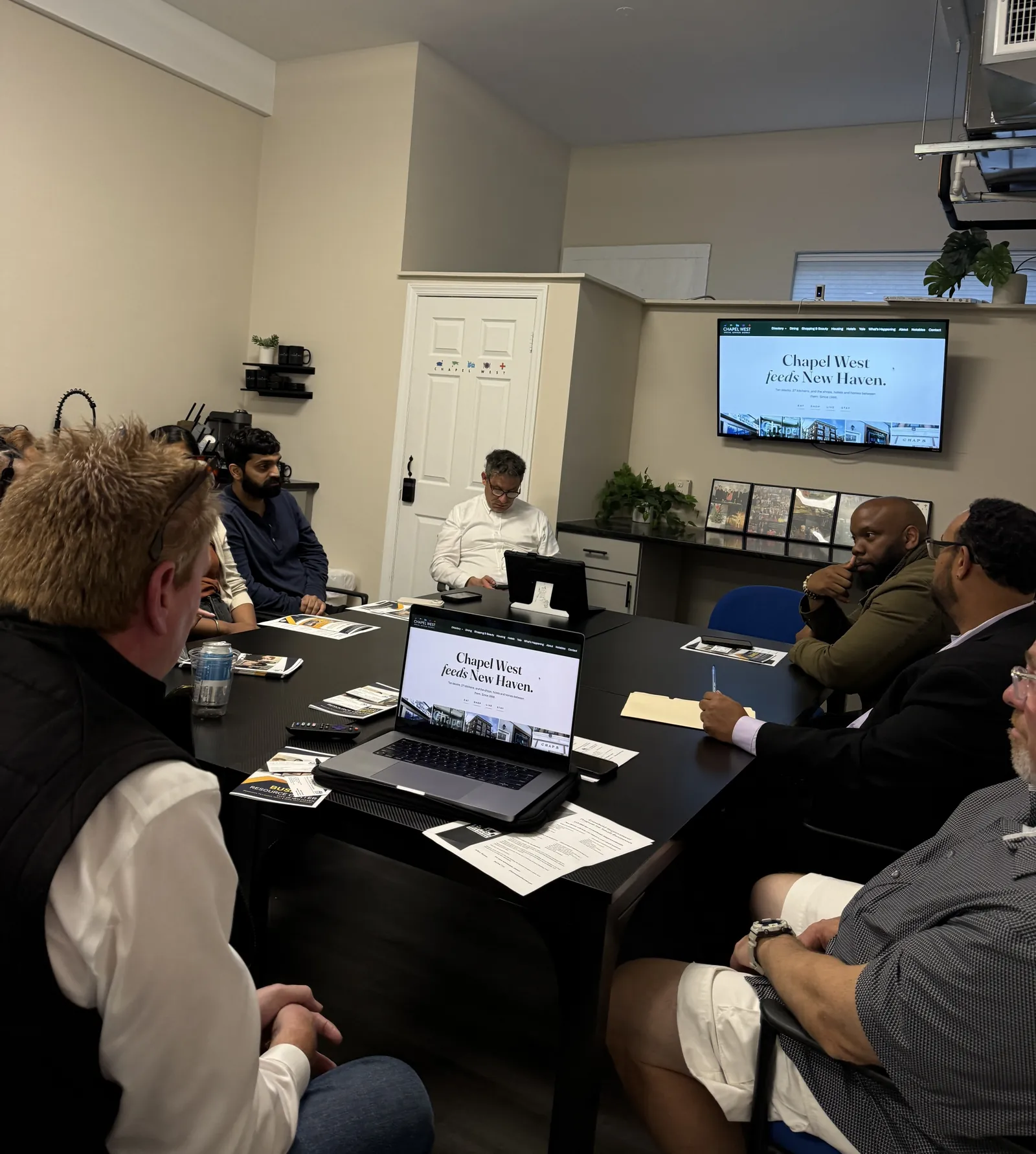 Business owners and City staff around the conference table at the Chapel West office, the Chapel West website on the wall screen and on a laptop, with City handouts spread on the table