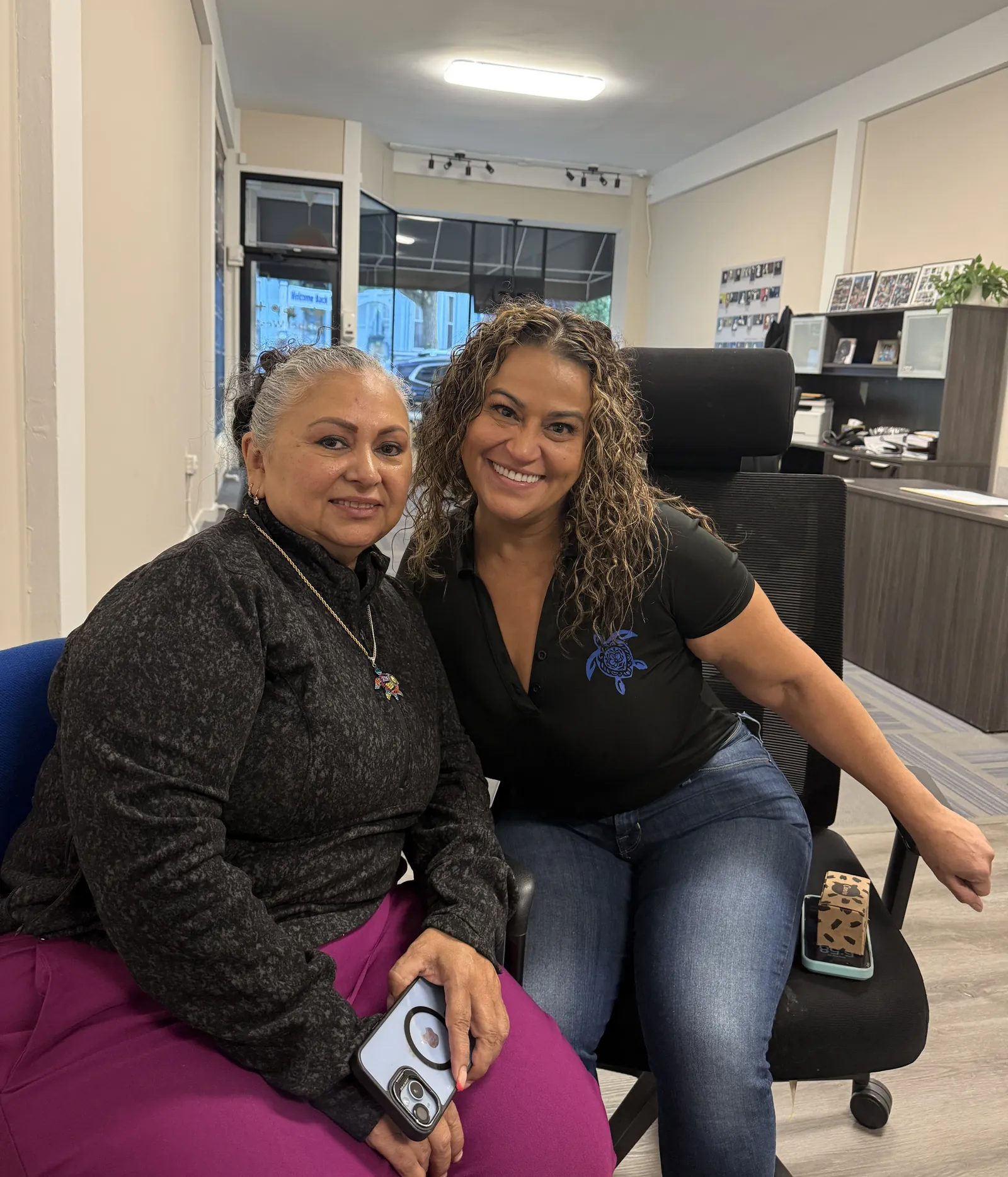 Two women smiling side by side in office chairs at the Chapel West office, one wearing a black shirt with a blue turtle logo