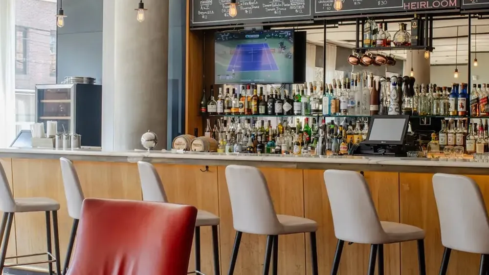 Inside Heirloom: a long bar with white stools, shelves of bottles and chalkboard menus, with a red leather armchair and lounge seating in front.