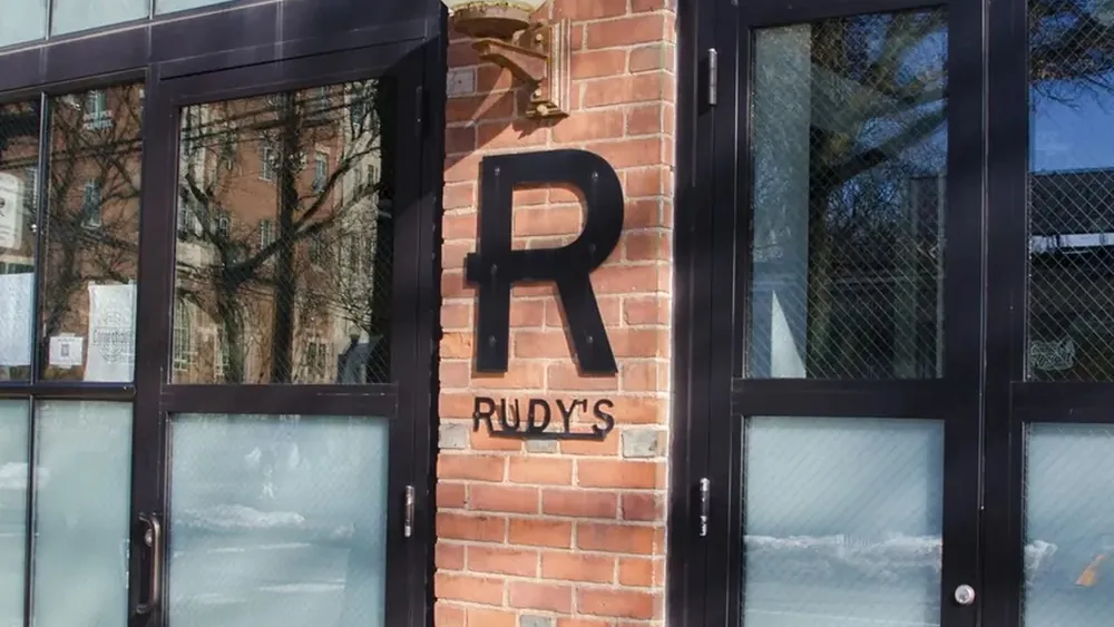 The front of Rudy's on a sunny day: brick wall, black-framed glass doors, a globe lamp and a large black R above the Rudy's name.