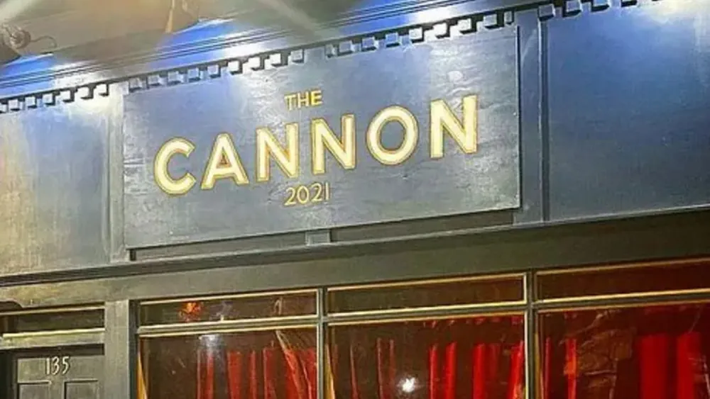 The Cannon at night: a dark green storefront with The Cannon 2021 in gold lettering, red curtains in the windows and a wooden railing out front.