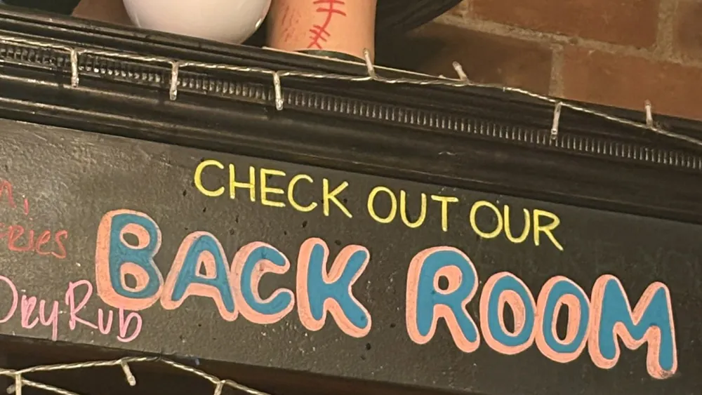 A chalkboard sign at Three Sheets framed in string lights, reading Check out our Back Room in yellow and blue chalk lettering.