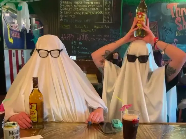 Two people dressed as ghosts in white sheets and sunglasses at the bar at Three Sheets, one holding a bottle on top of their head, under the chalkboard drafts list.