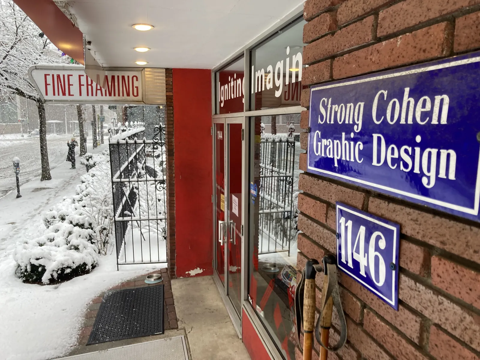 The blue enamel Strong Cohen Graphic Design sign and a 1146 number plate on the brick front of the Chapel Street storefront, beside Hull's fine framing entrance, in falling snow
