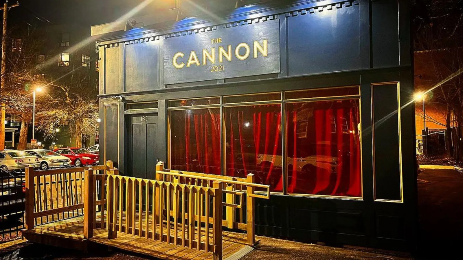 The Cannon lit at night, gold lettering on a black facade with red curtains in the windows and a wooden deck in front