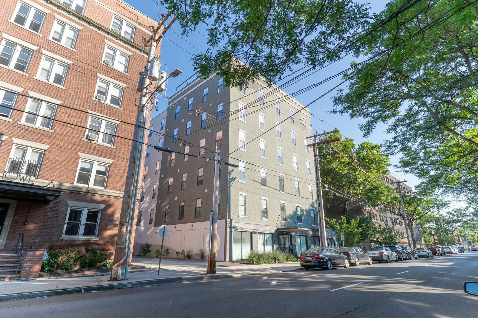 The Elm seen from across Howe Street, a modern grey and olive brick building with a pale ground floor rising beside the older red brick apartments next door, under a clear sky